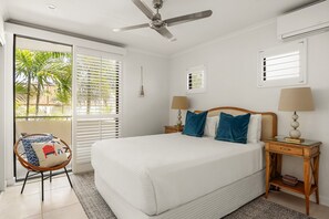 Une chambre à coucher avec un lit, deux tables de chevet avec des lampes, un ventilateur de plafond et une vue sur les palmiers par la fenêtre.