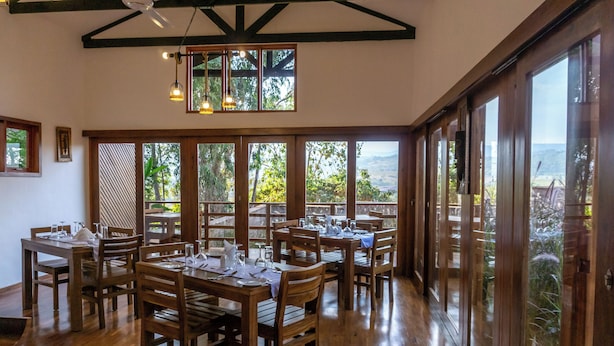 A restaurant with wooden tables and chairs, large windows with a view of the outdoors, and hanging pendant lights.