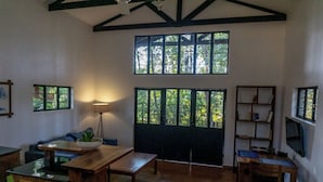 A spacious living room with a high ceiling, large windows, a wooden dining table, a sofa, and a TV mounted on the wall.