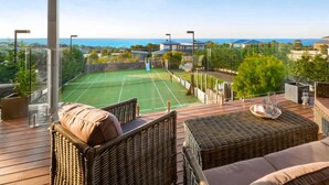 Un court de tennis avec un filet, entouré d’une terrasse en bois agrémentée de meubles en osier et d’une rambarde en verre.