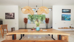 Un vaste espace dédié à la salle à manger, avec une grande table en bois, deux luminaires suspendus en osier et une vue sur la verdure environnante.