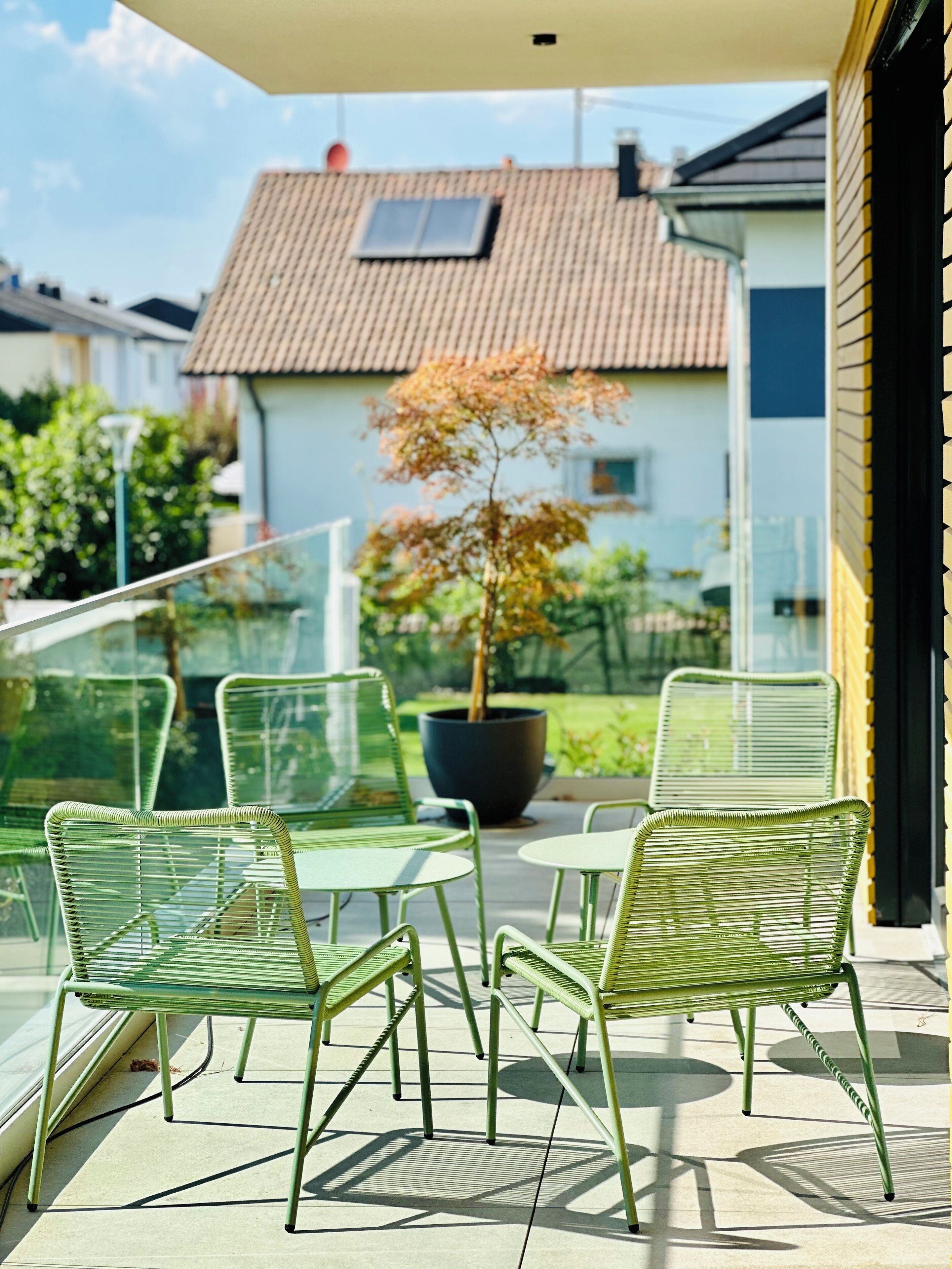 Eine Terrasse mit grünen Metallstühlen und einem Tisch, ein Topfbaum und im Hintergrund ein Haus mit einem Ziegeldach.