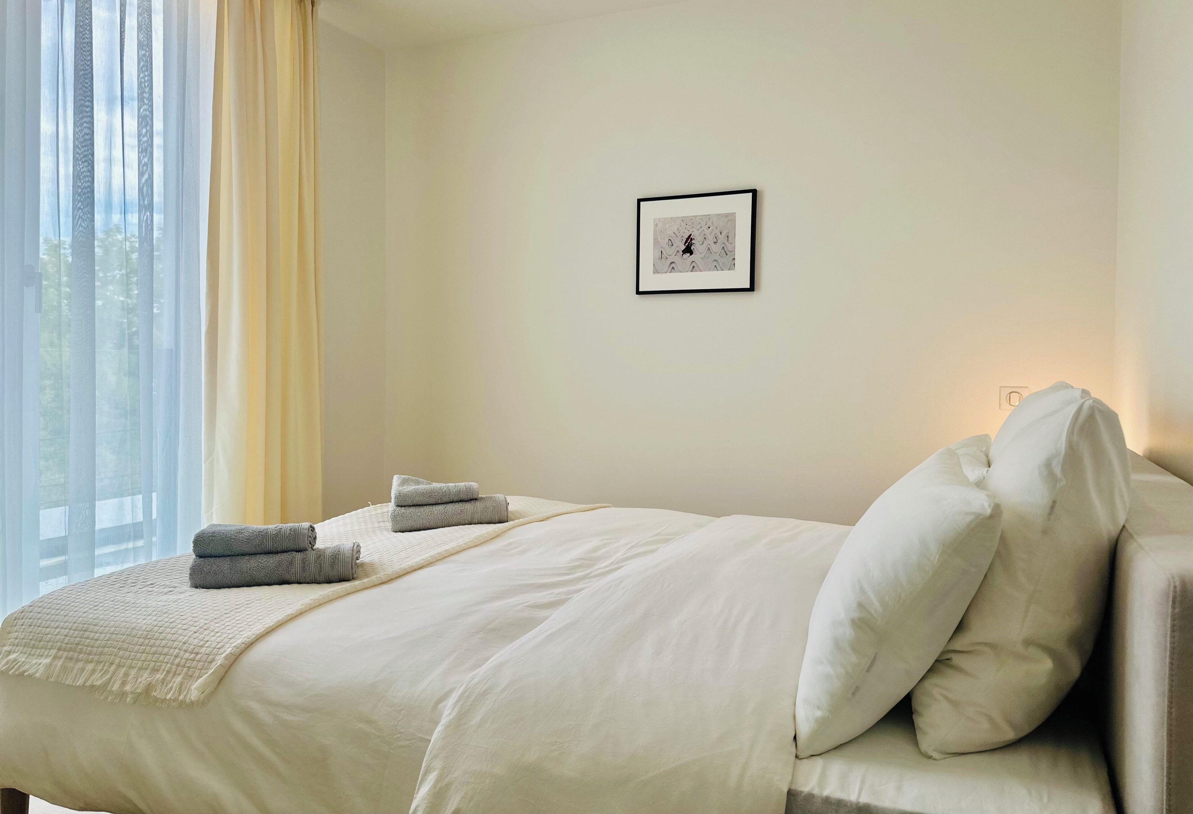 Een keurig opgemaakt bed met kussens en gevouwen handdoeken, een ingelijst schilderij aan de muur en een raam met gordijnen.