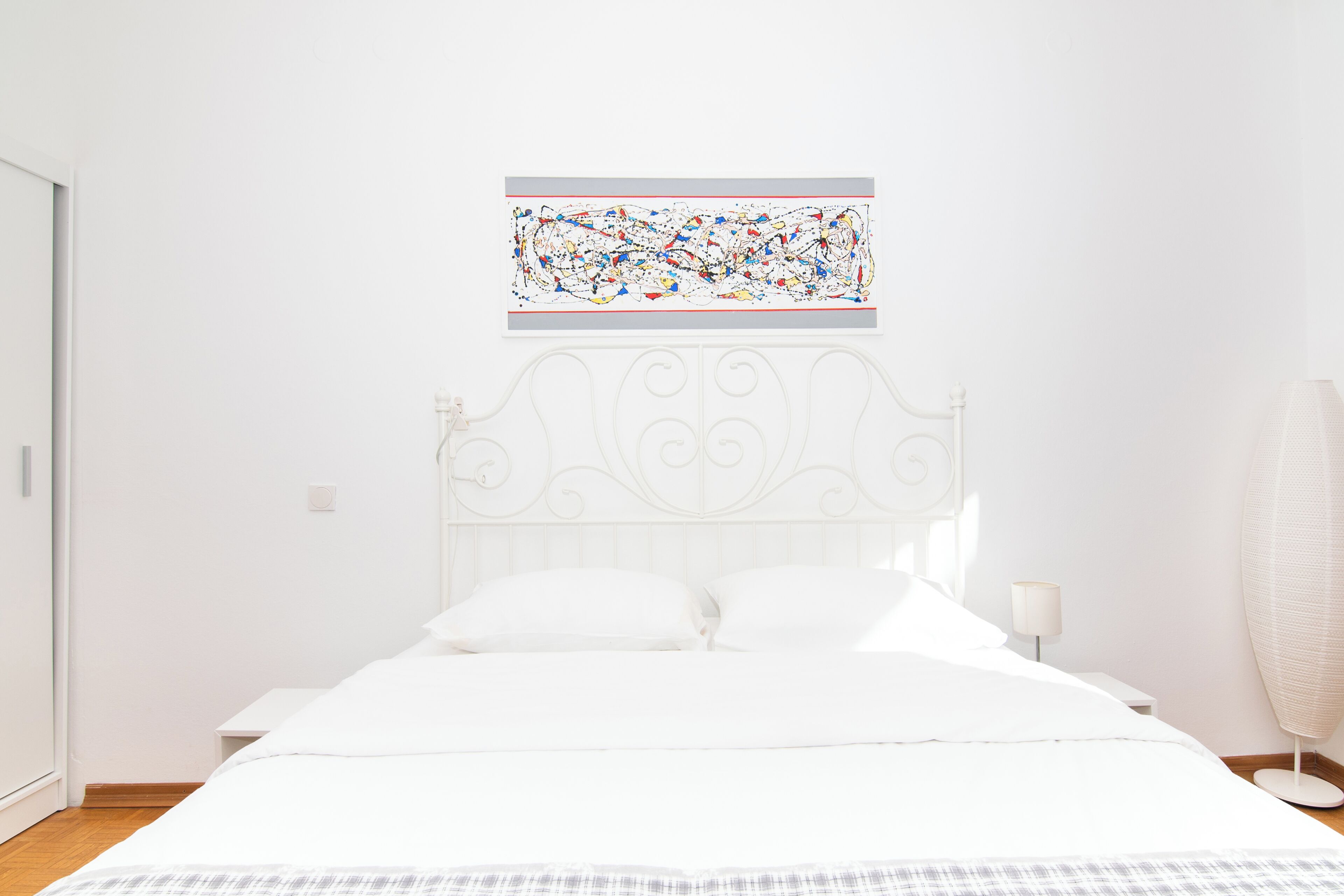 Ein Schlafzimmer mit einem weißen Bett, einem dekorativen Kopfteil, einem an der Wand hängenden abstrakten Gemälde, einem Nachttisch mit Lampe und einer Stehlampe.