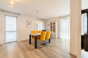 A modern dining area with a wooden table and yellow chairs, large windows with sheer curtains, and a built-in cabinet with glass doors. - exklusives Appartement in Rechlin (Rechlin)