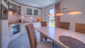 Une cuisine moderne avec des armoires blanches, un four encastré et un coin repas agrémenté de chaises et d’une table en osier.