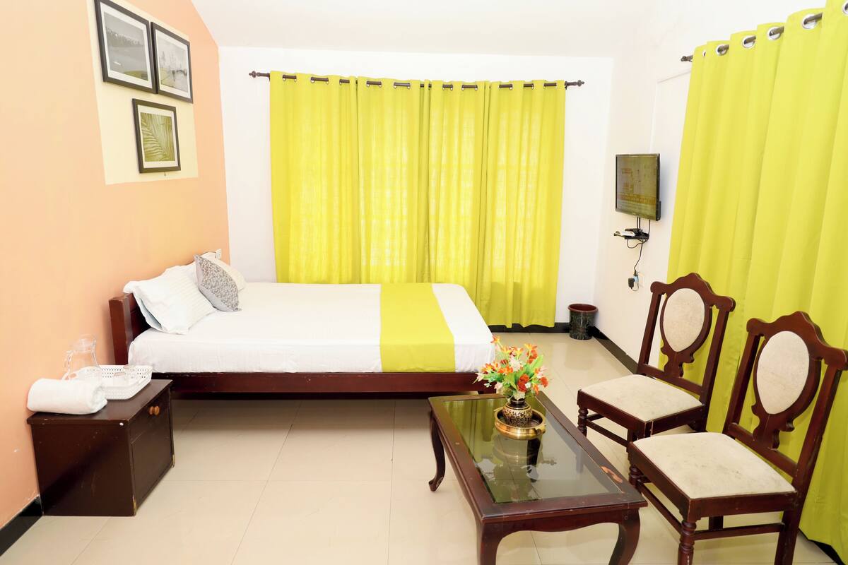 A hotel room with a bed, a small table with a vase of flowers, two wooden chairs, a nightstand, and a television on the wall.