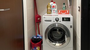 Een kleine wasruimte met een wasmachine, een dweil, schoonmaakspullen en een rode emmer.