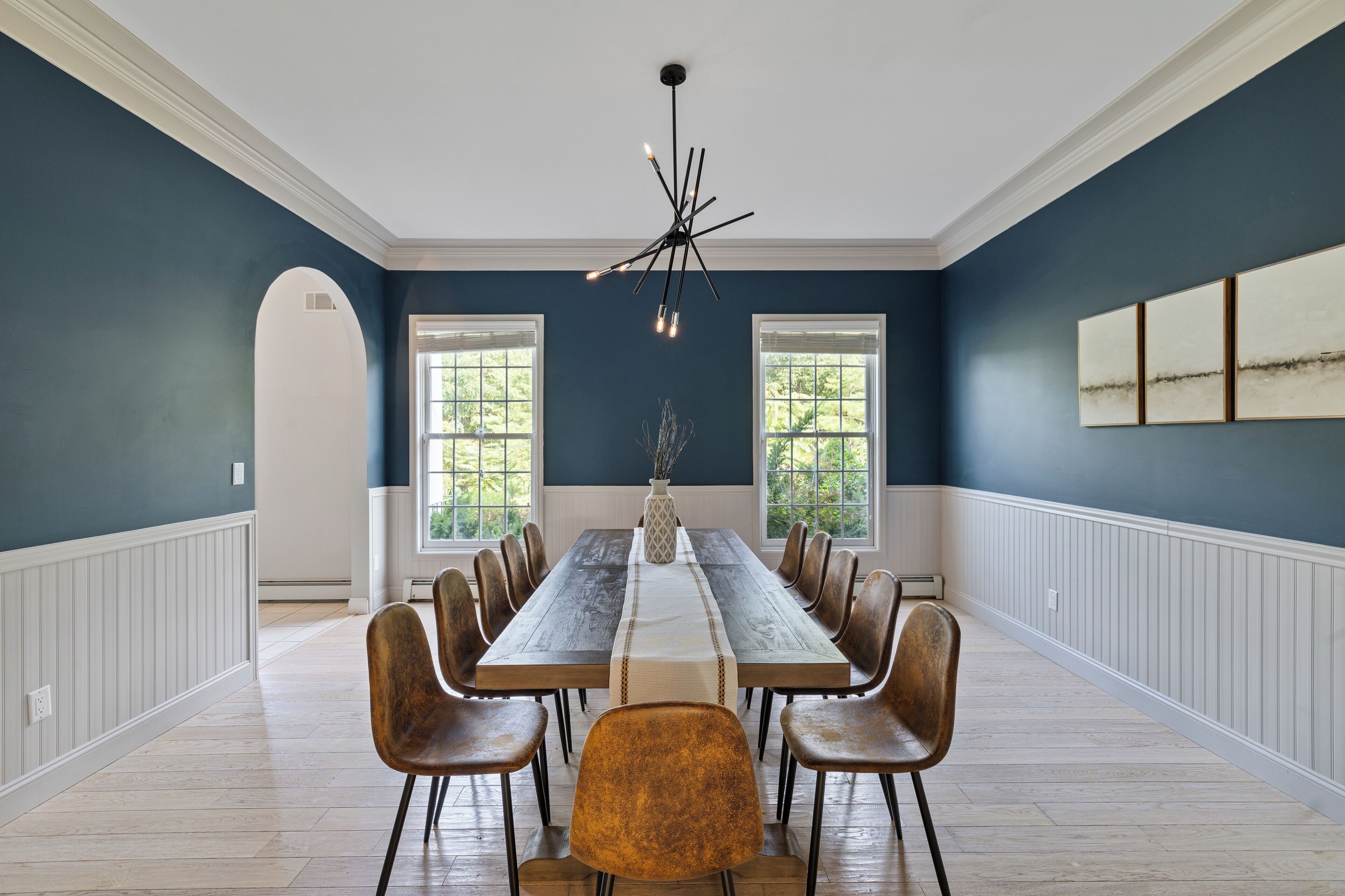 Une salle à manger avec une longue table, des chaises, un lustre et des fenêtres.