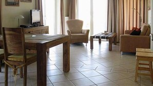 Uma sala de estar com uma mesa de jantar de madeira, uma poltrona bege, um sofá e uma televisão sobre um móvel.