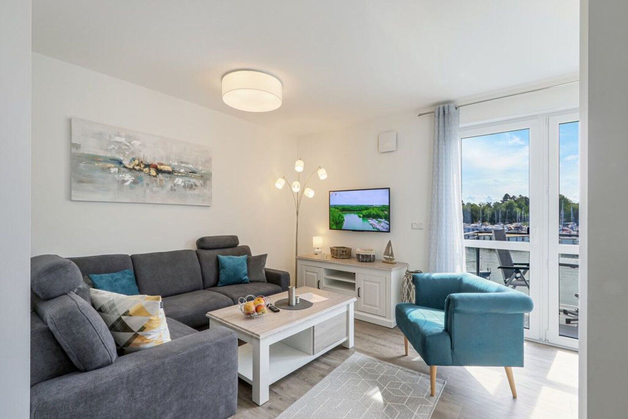 A modern living room with a grey sofa, a blue armchair, a coffee table, and a flat-screen TV. There is a painting on the wall and a view of the outdoors through large windows.