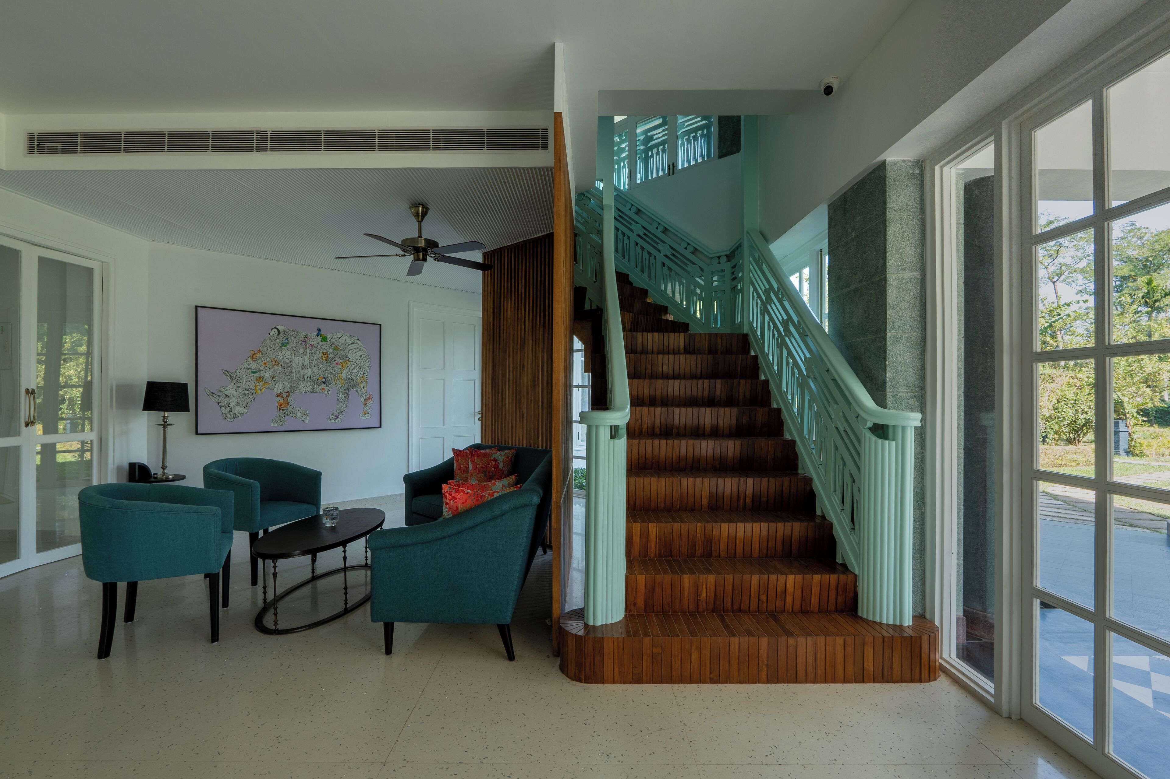 A spacious living area with teal armchairs, a wooden staircase with teal railings, and a large window offering a view of the outdoors.