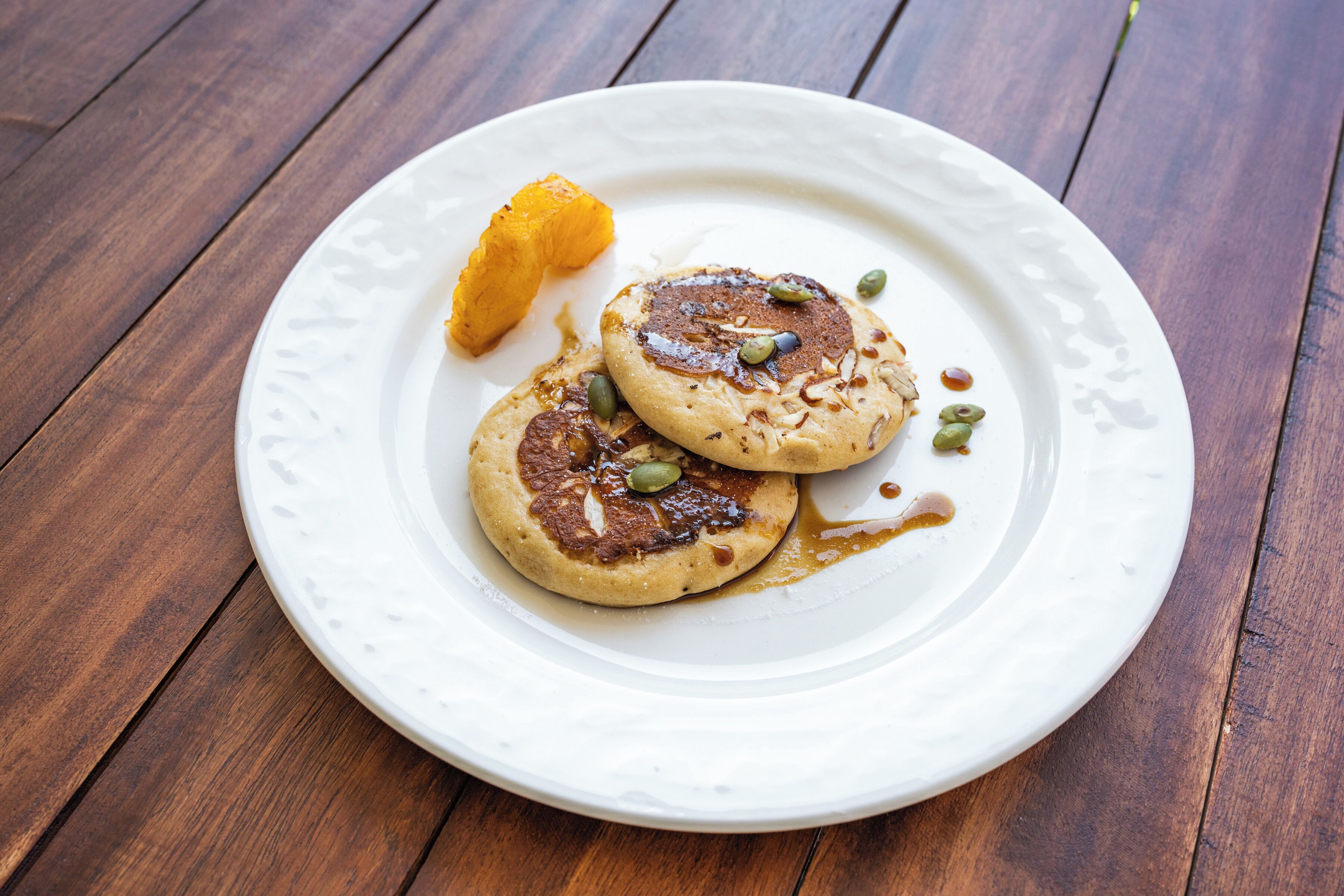 A white plate with two pancakes, a slice of orange, and some seeds on a wooden table.