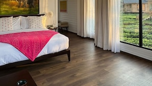 Un'ampia camera da letto con un grande letto, una ventola a soffitto, un dipinto di elefanti e vista su una casa attraverso ampie finestre.