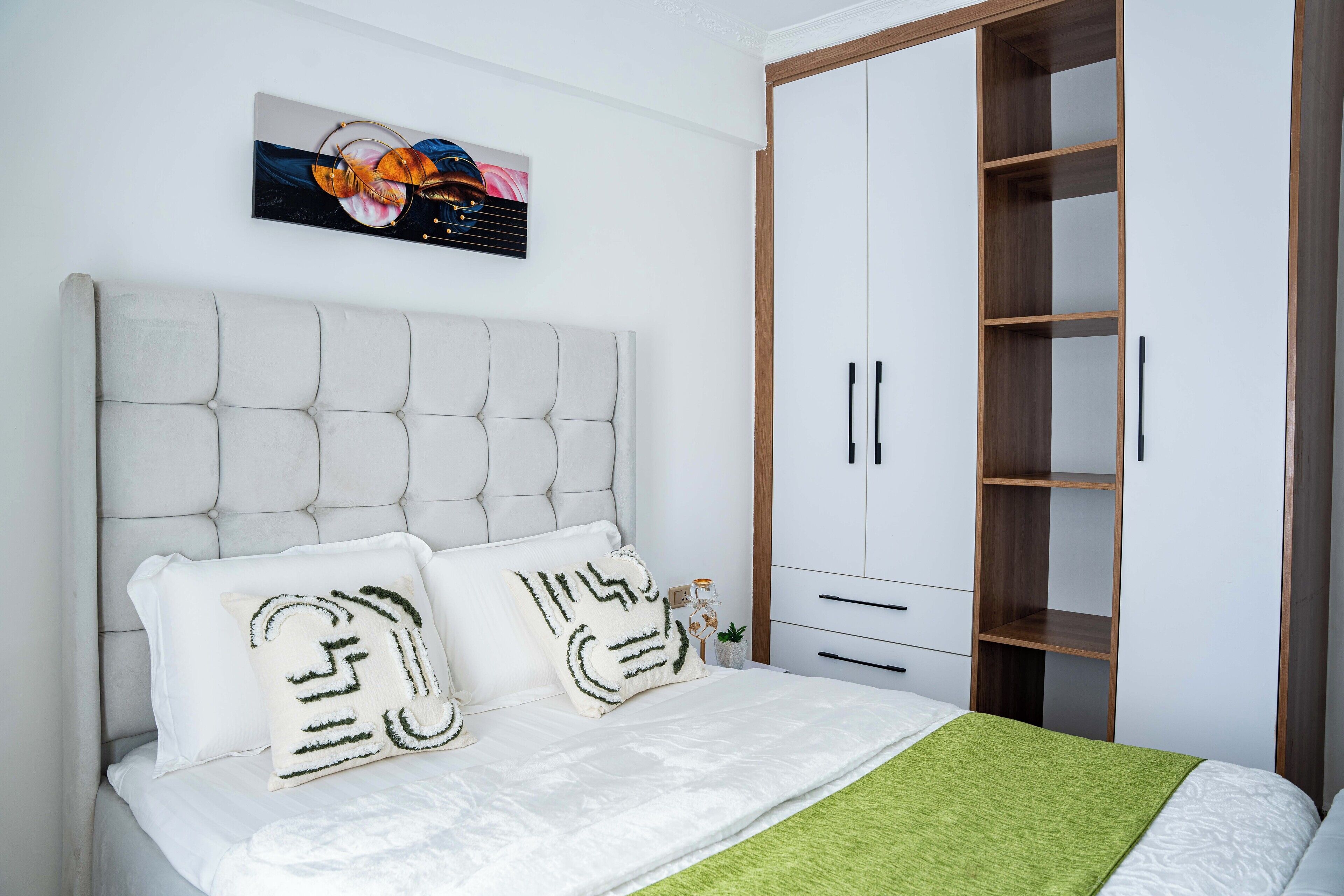 A bedroom with a tufted headboard, two decorative pillows, a green throw blanket, a white bedspread, a wooden wardrobe, and a framed abstract painting on the wall.