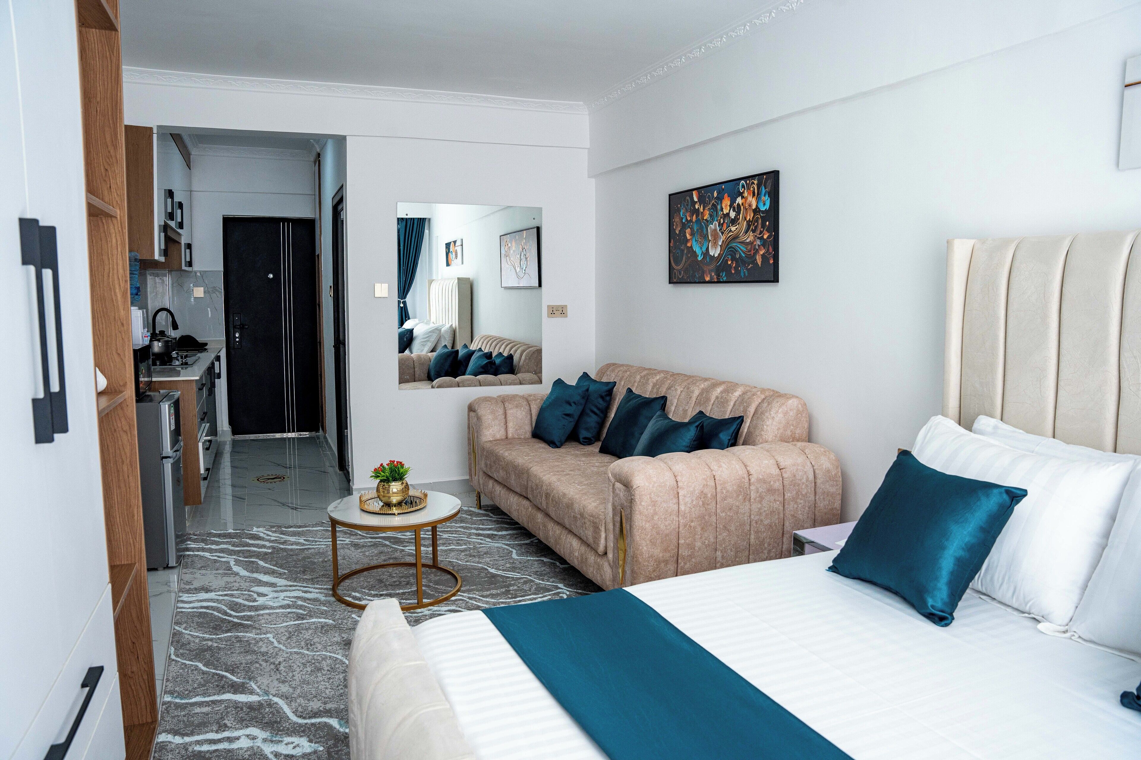 A modern hotel room with a large bed, a sofa, a small round table, and a kitchen area in the background.