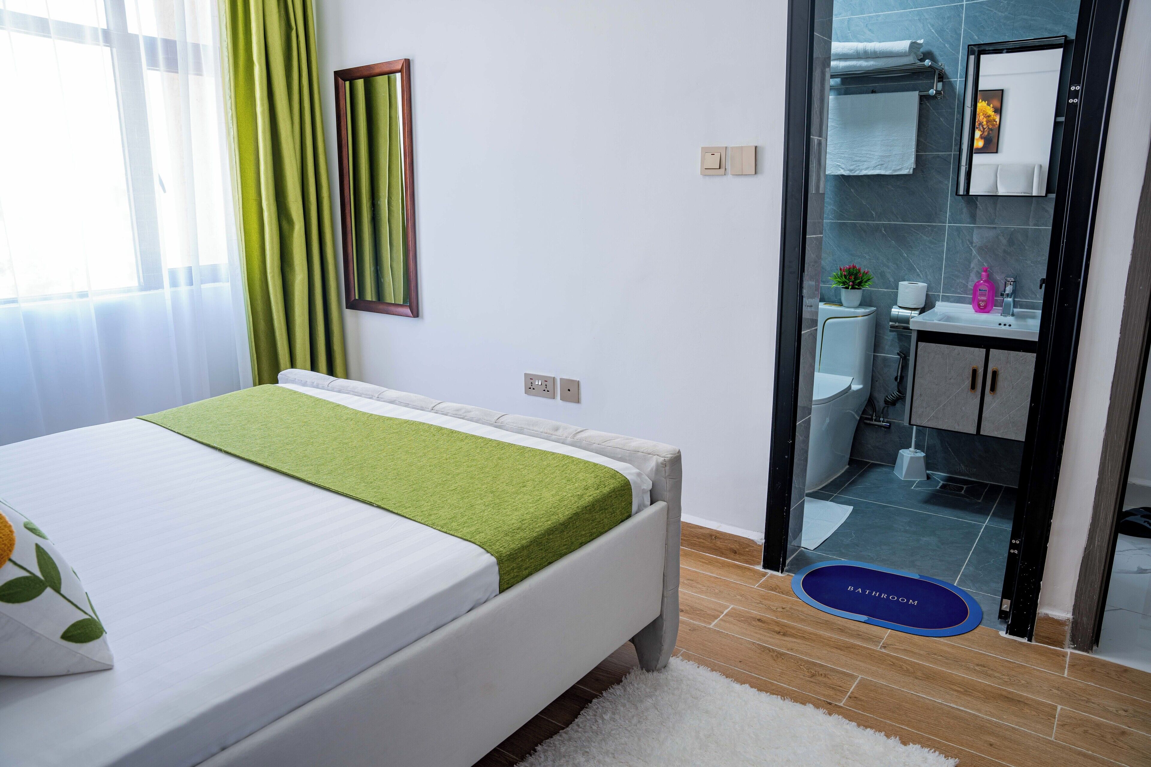 A modern bedroom with a bed, a green throw blanket, a white pillow, and a bathroom visible through an open door.