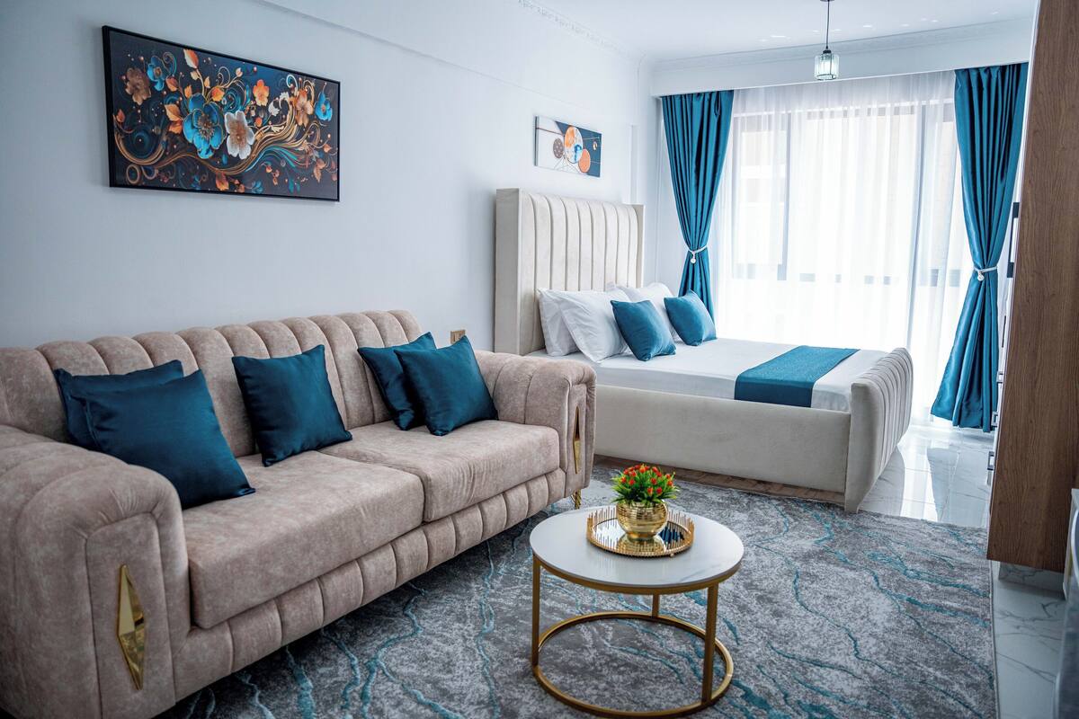 Quarto de hotel moderno com sofá bege, cama branca e mesa de centro redonda com uma tigela decorativa.