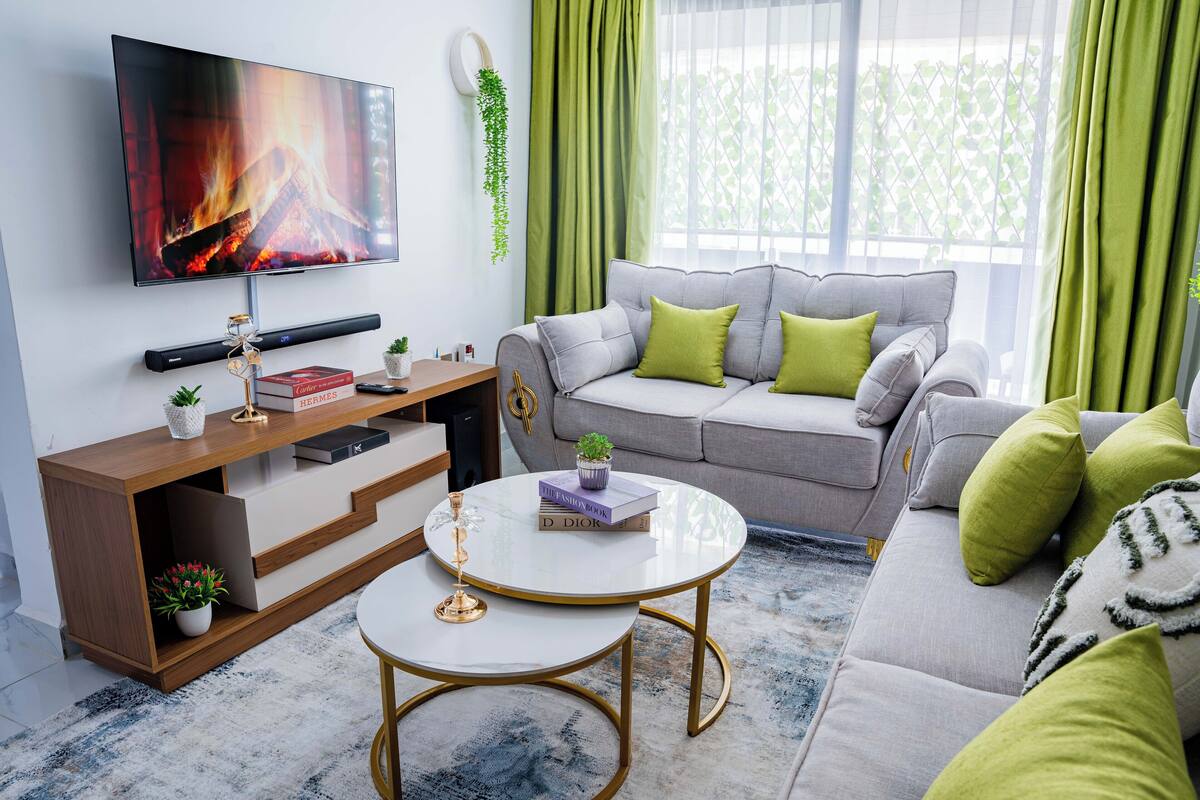 Uma sala de estar moderna com um sofá cinzento, almofadas verdes, uma televisão de tela plana, uma mesa de centro e uma estante.