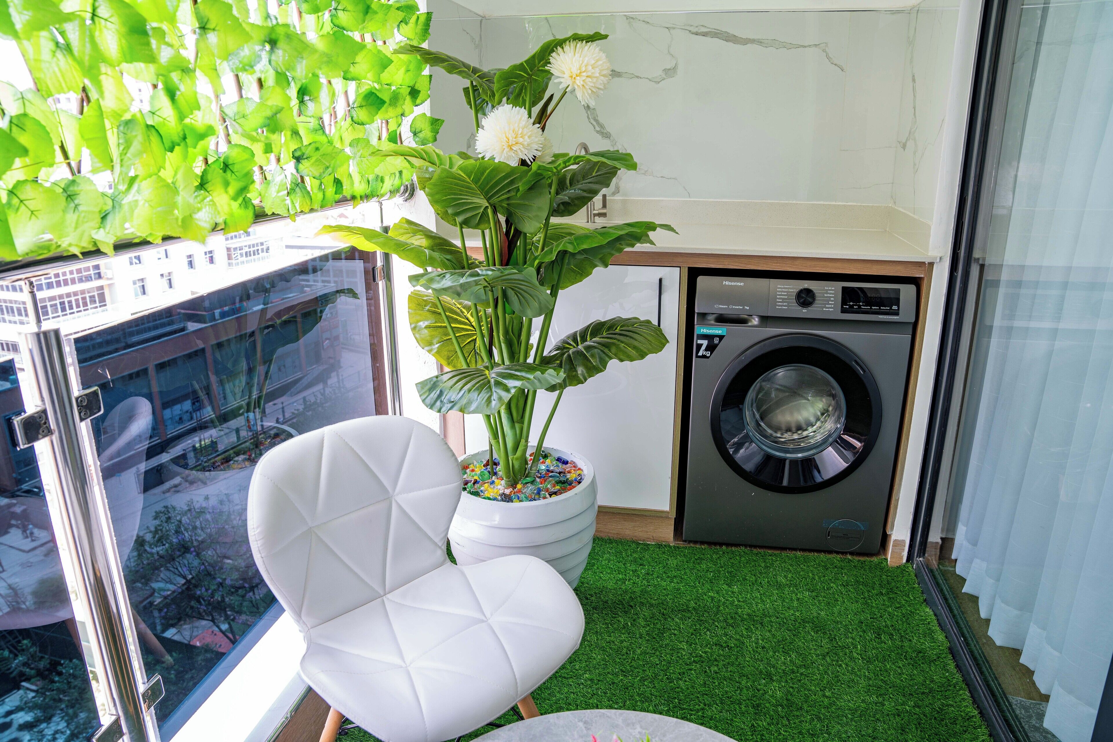 Une salle de bain moderne avec une chaise blanche, une grande plante en pot, un lave-linge et une vue à travers une porte vitrée.