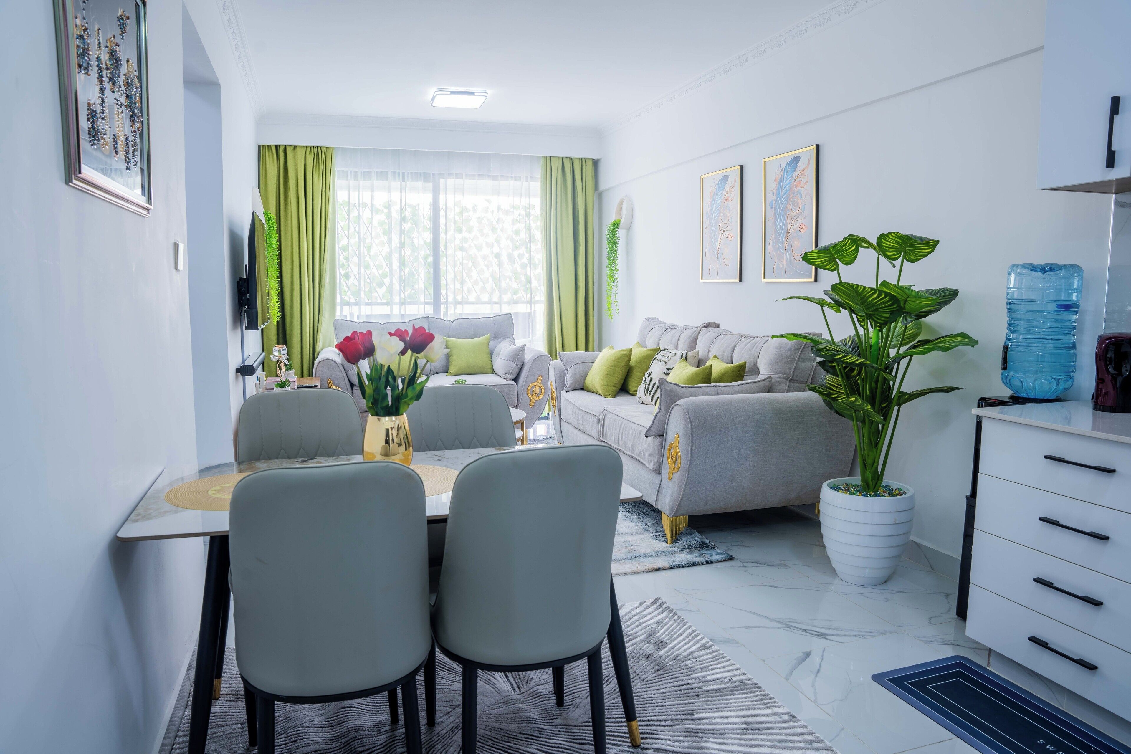 A modern living room with a grey sofa, a dining table with chairs, a large potted plant, and artwork on the walls.