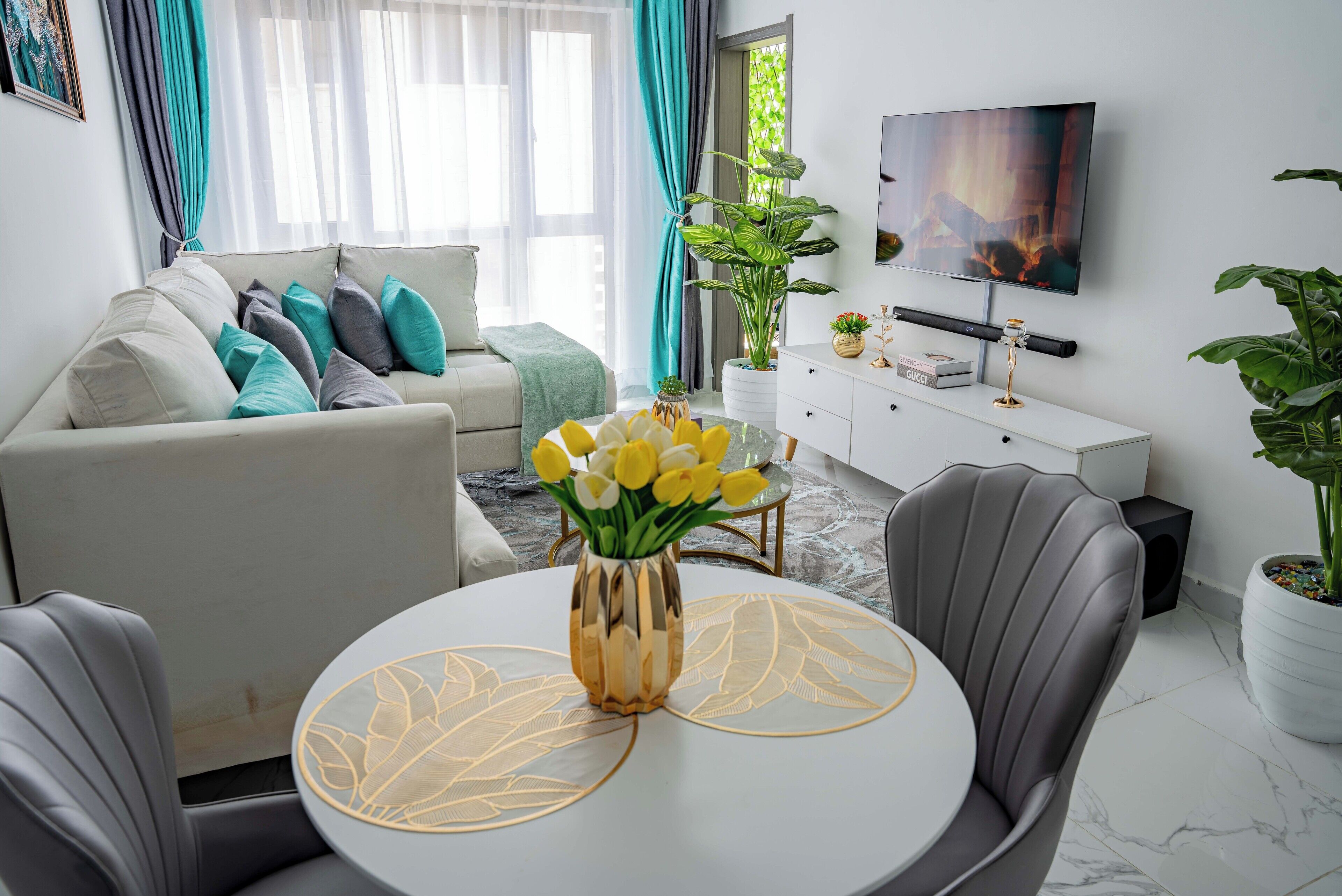 A modern living room with a sofa, a dining table with a vase of yellow flowers, a TV on the wall, and a potted plant.
