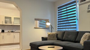 A modern living room with a grey sofa, a small round table, and a lamp. There is a kitchen area with cabinets and a countertop visible through an archway. - Stylish apartment in a central location (Dissen am Teutoburger Wald)