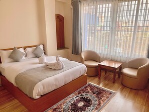 A hotel room with a large bed, two armchairs, a small table, and a view of the outside through sheer curtains.
