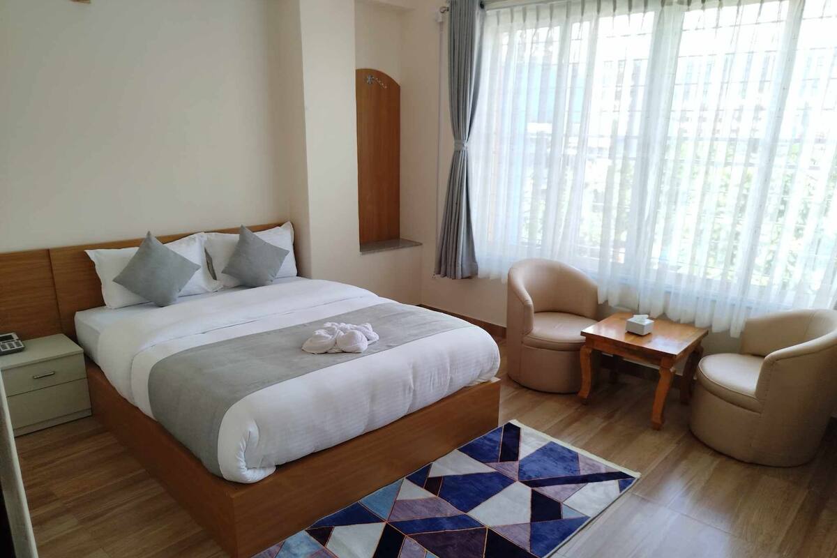 Una habitación de hotel con cama, dos sillas, una mesita, una mesita de noche y una ventana con cortinas.