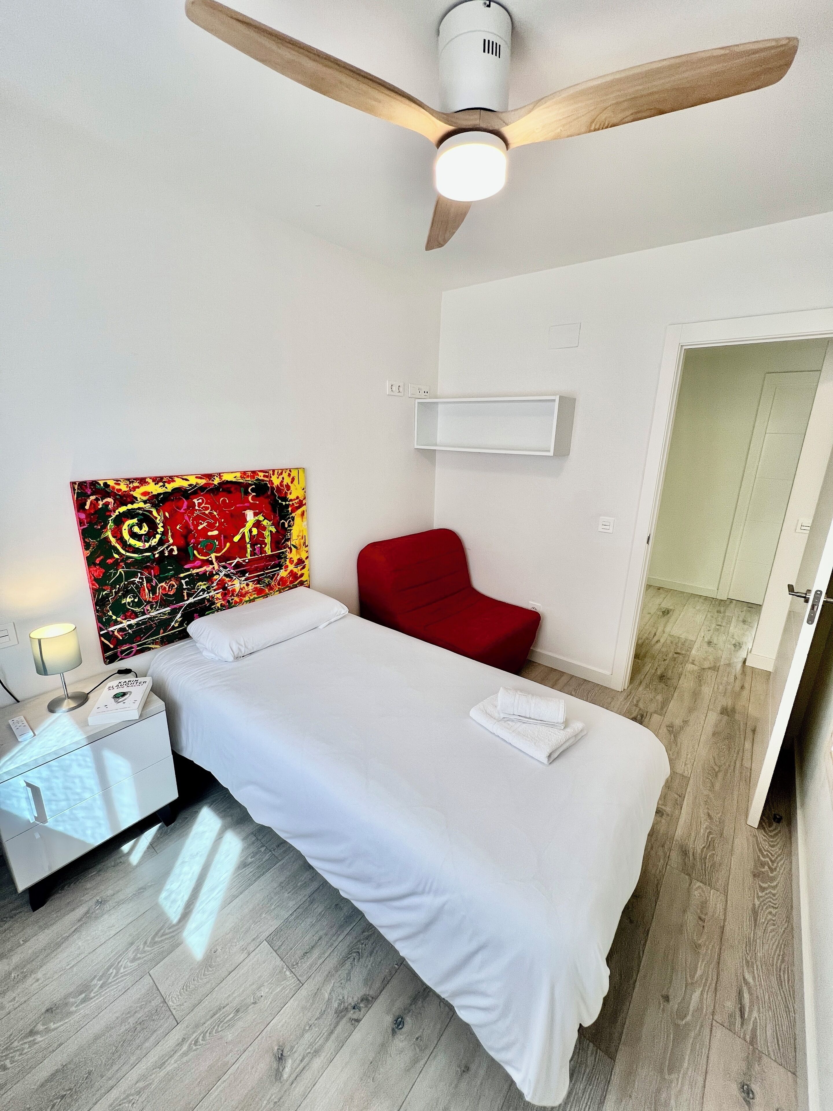 Habitación de hotel con cama, una silla roja, ventilador de techo, mesita de noche con lámpara y estante en la pared.