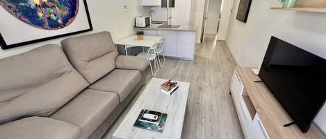 Un salon moderne avec un canapé beige, une table basse sur laquelle repose un livre, un téléviseur à écran plat et une cuisine en arrière-plan.