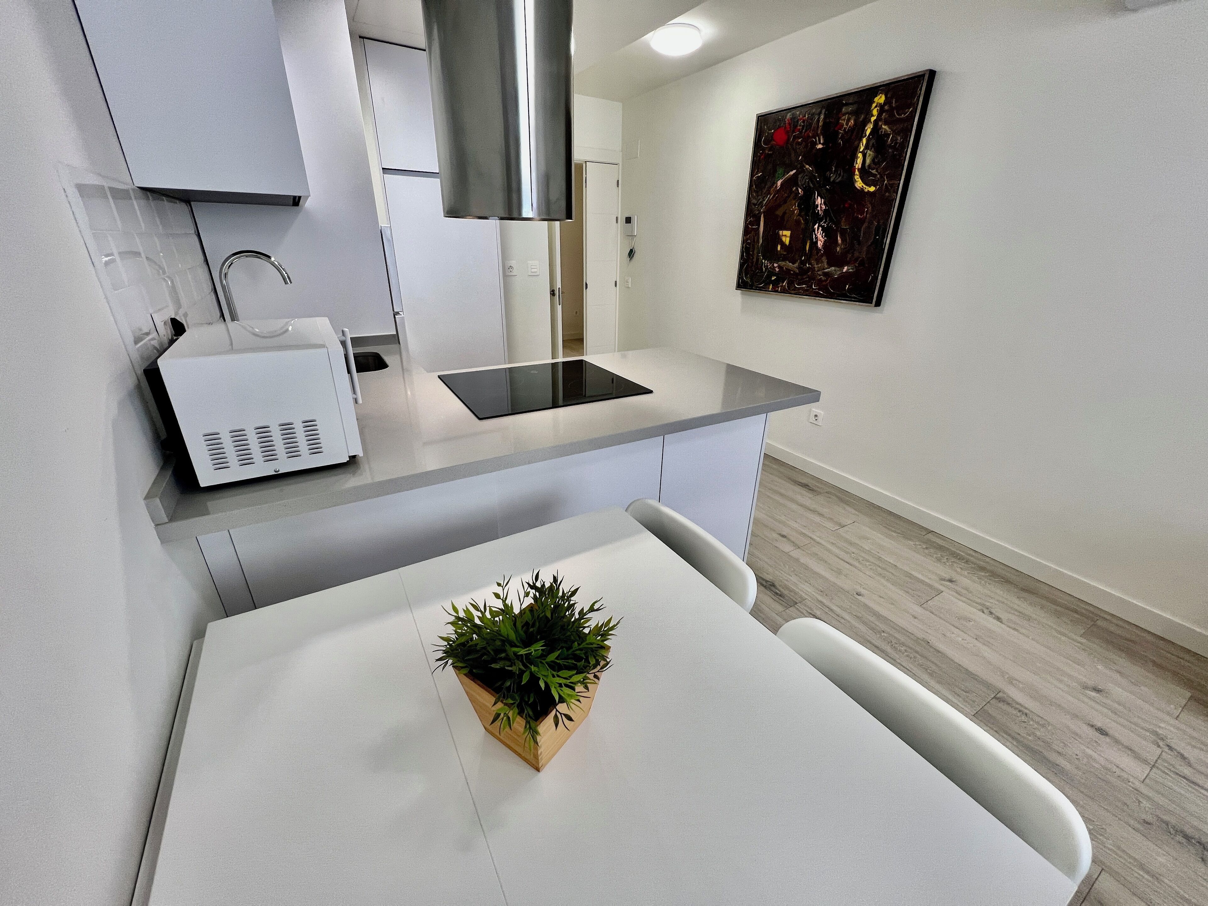 A modern kitchen with white cabinetry, a built-in microwave, a stainless steel range hood, and a small dining table with a potted plant.