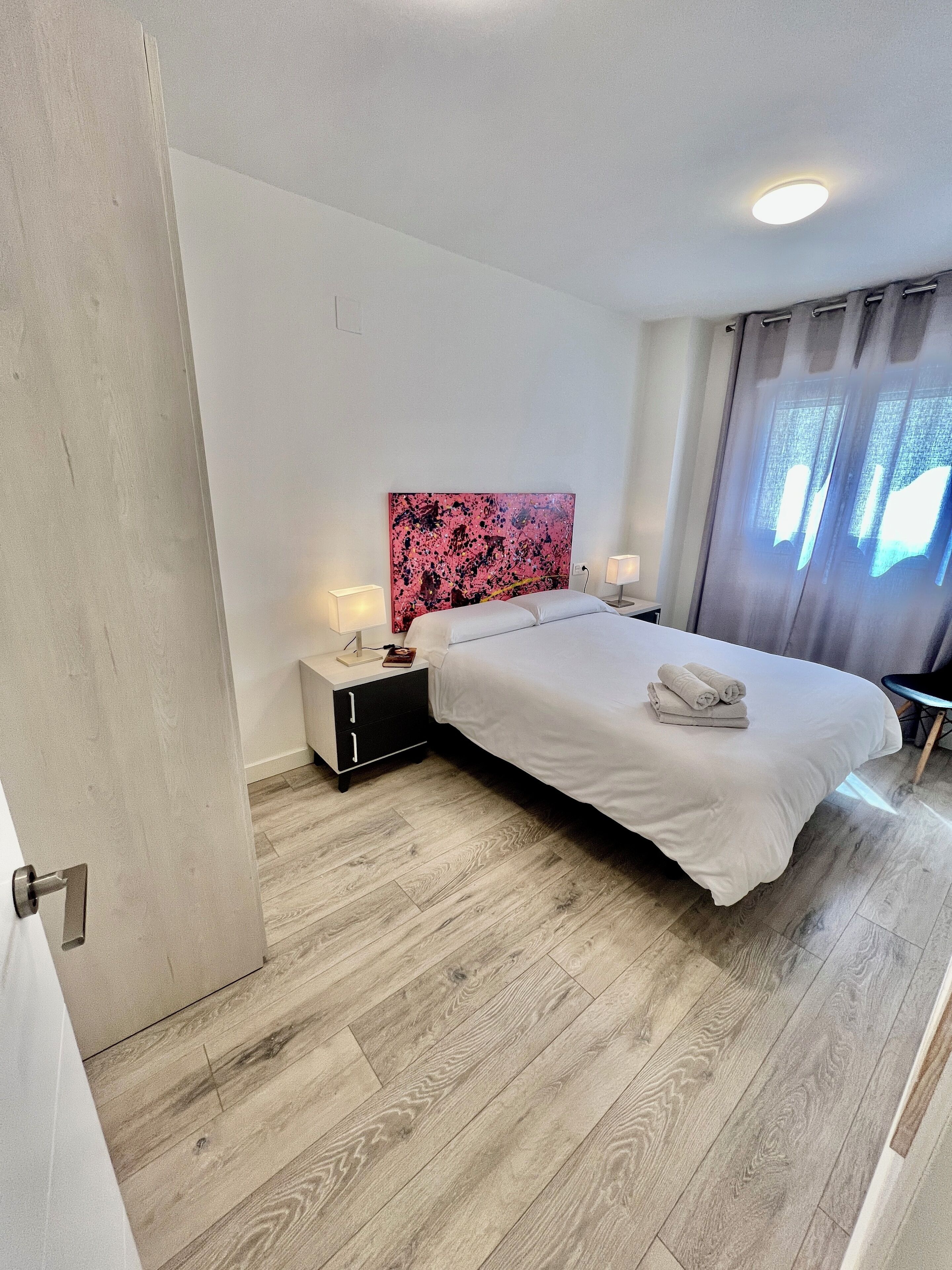 Ein modernes Schlafzimmer mit Holzboden, einem Bett mit weißer Bettwäsche, einem Nachttisch mit Lampe und einem großen abstrakten Gemälde an der Wand.