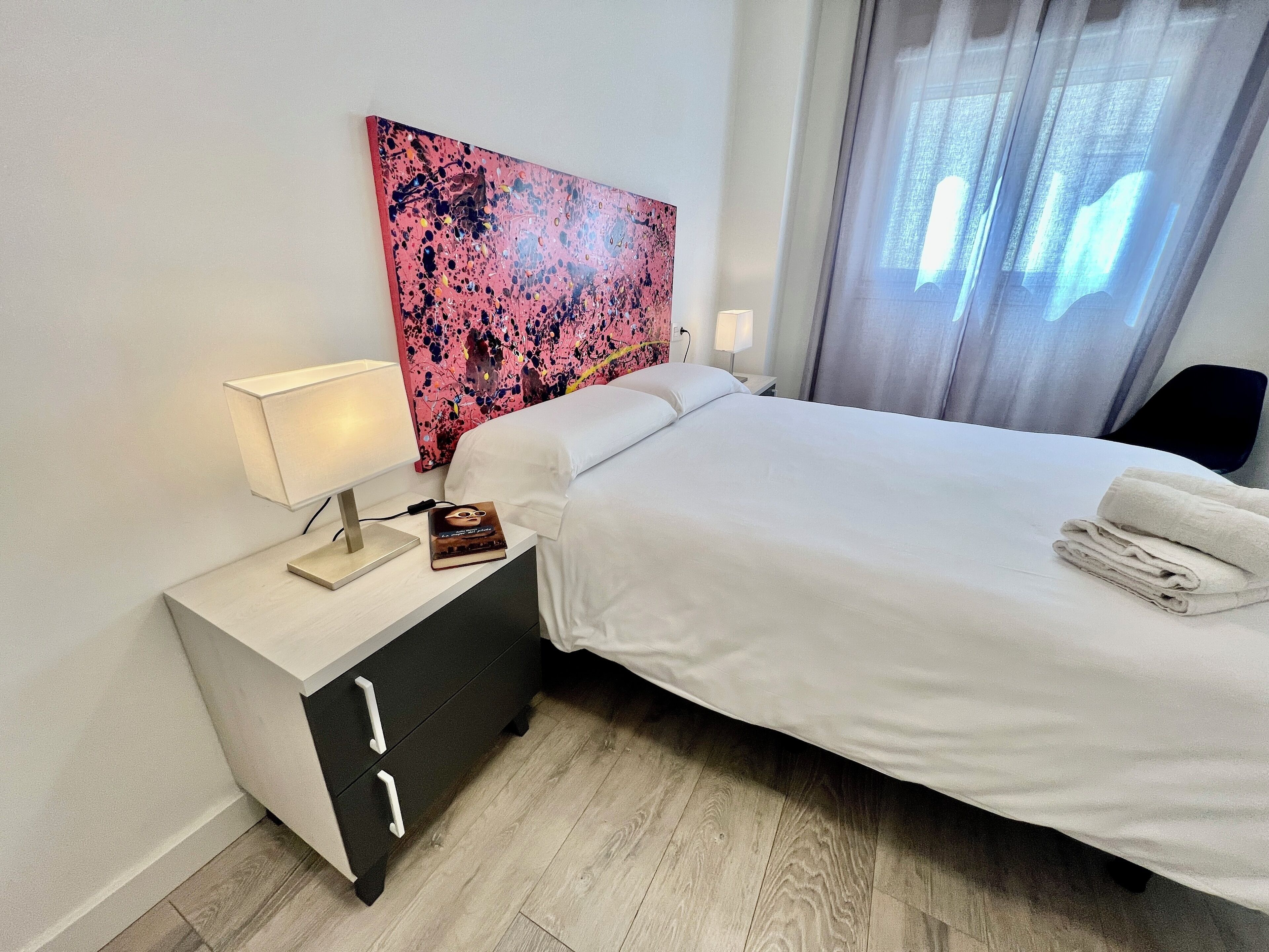 A neatly made bed with white linens, a bedside lamp, a book, and a folded towel on a nightstand.