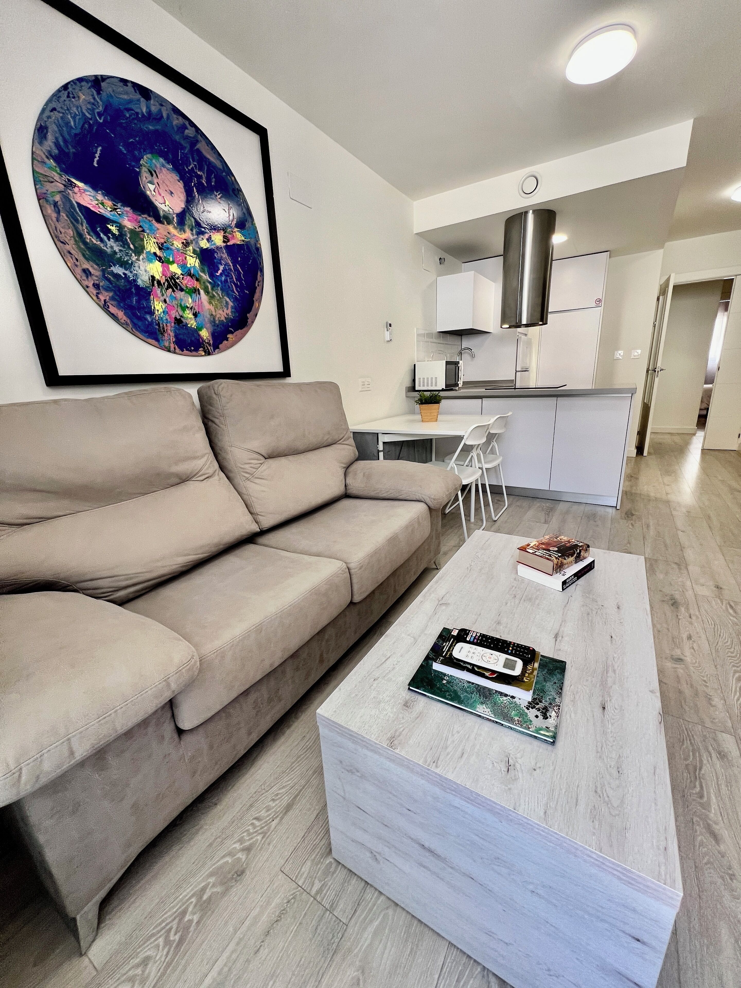 A modern living room with a beige sofa, a coffee table with books, a kitchen in the background, and a large abstract painting on the wall.