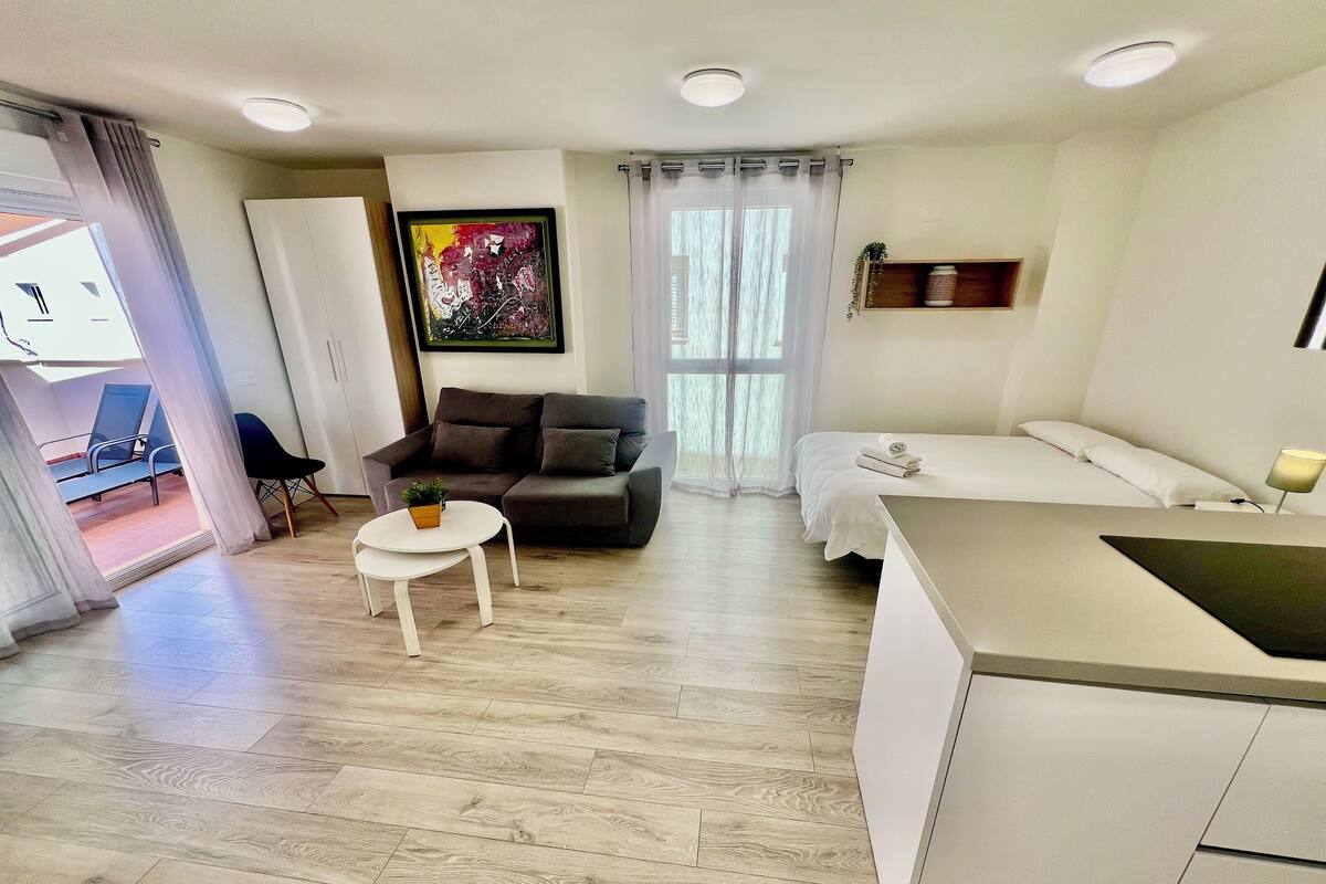 Una sala de estar moderna y bien iluminada con sofá, una mesita redonda, cama y zona de cocina.