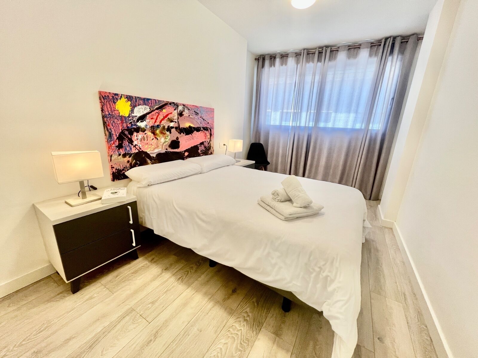 A hotel room with a bed, a bedside table with a lamp, a painting on the wall, and a window with curtains.