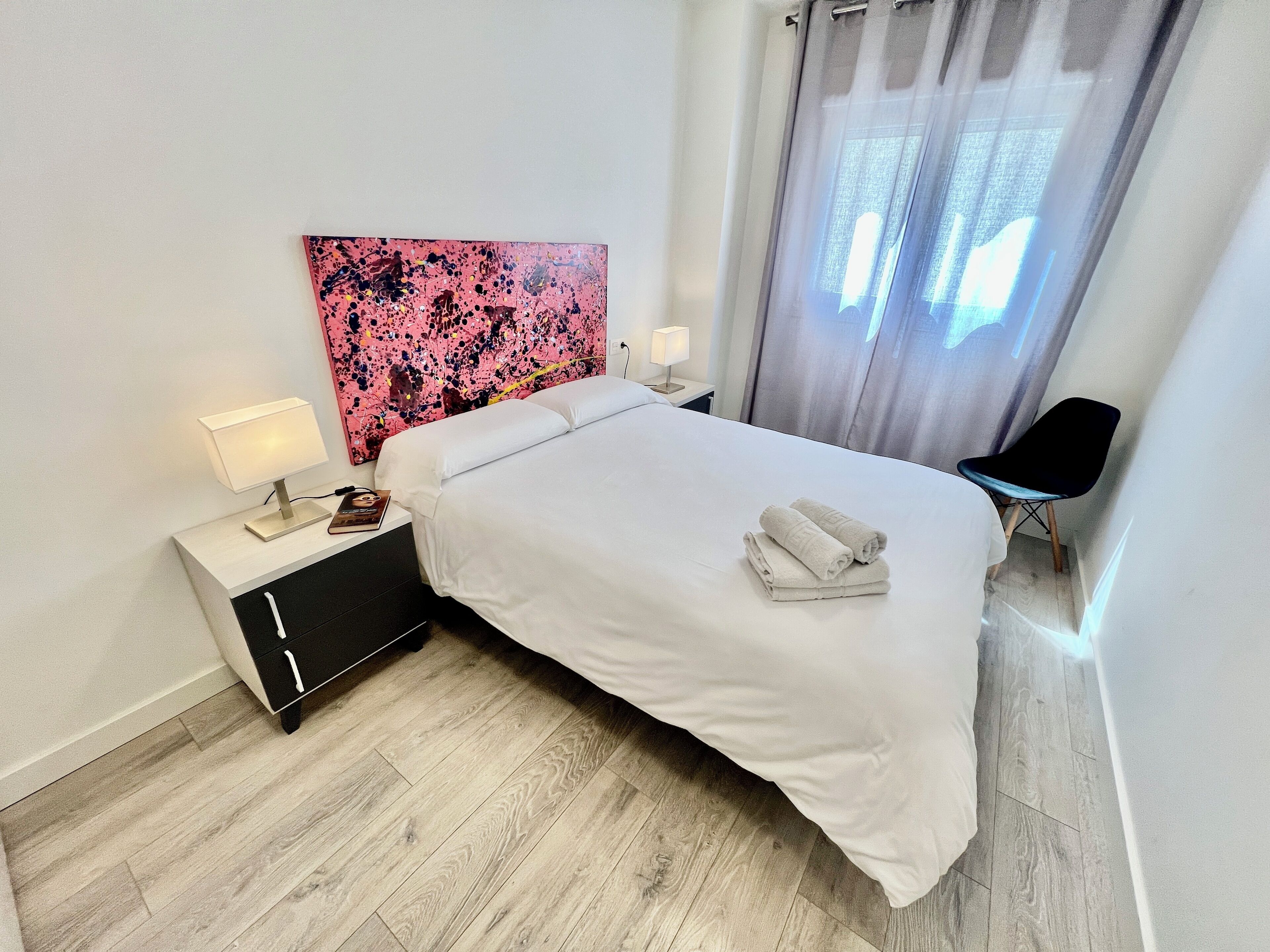 A neatly made bed with white linens and two folded towels, a bedside table with a lamp and a book, a black chair, and a large abstract painting on the wall.