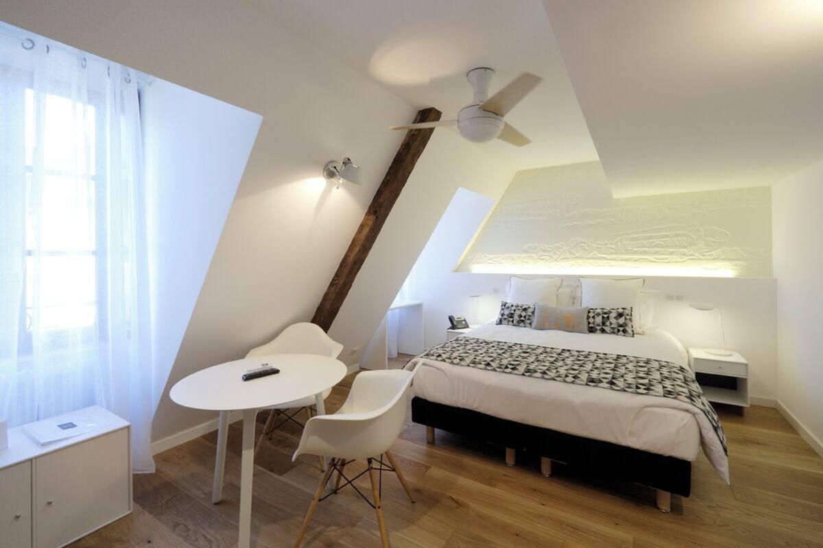 A modern bedroom with a sloped ceiling, a bed with patterned bedding, a bedside table, a nightstand, a desk with a chair, and a window with sheer curtains.