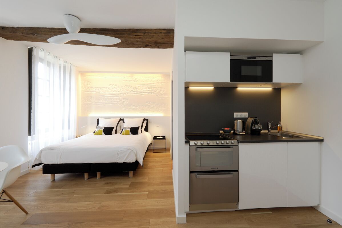 A modern hotel room with a bed, bedside table, and a kitchenette with a stove and oven.
