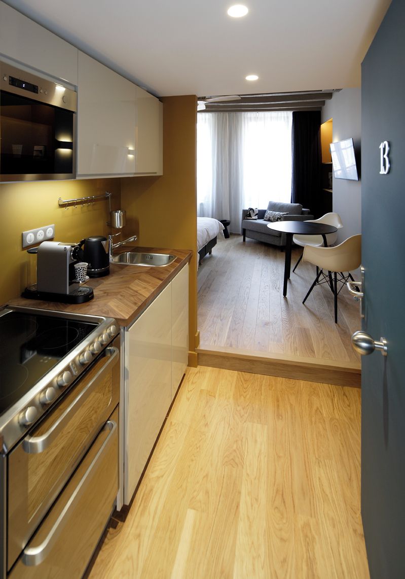 Une cuisine compacte dotée d’appareils électroménagers modernes, un coin repas avec une petite table et des chaises, et une vue sur une chambre avec un lit et une fenêtre.