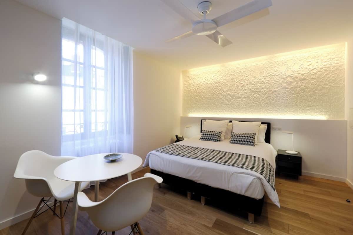 A modern hotel room with a bed, a ceiling fan, a small table with chairs, and a large window with curtains.