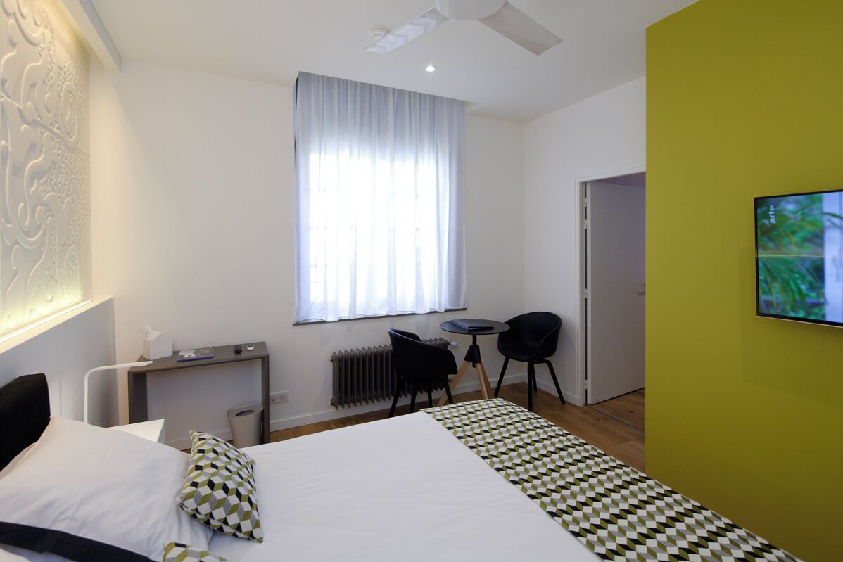 A hotel room with a bed, a desk, a chair, a radiator, a television, and a window with curtains.