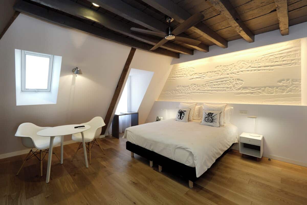 A bedroom with a bed, bedside tables, a desk, and chairs. There is a window and a ceiling fan.