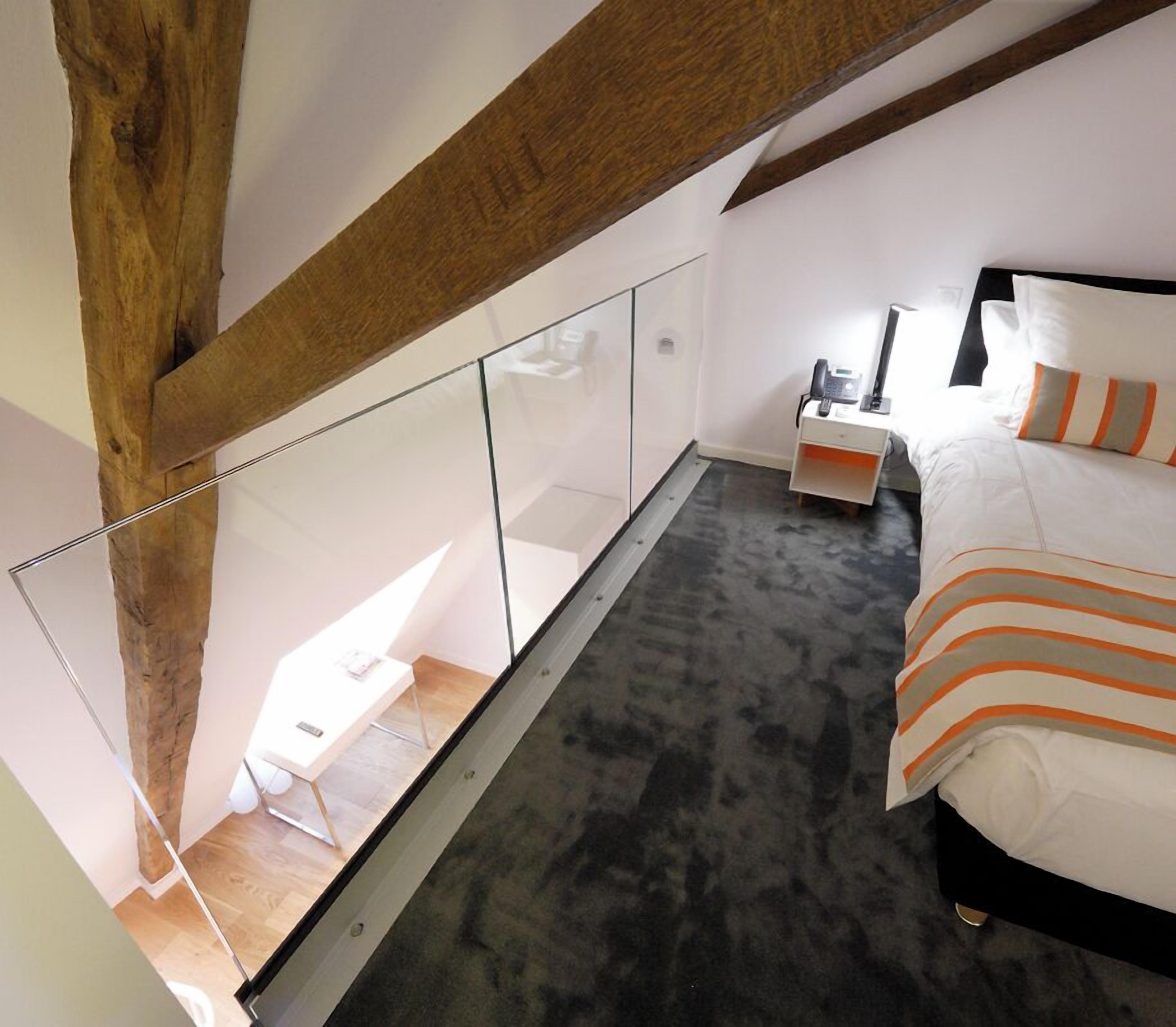 A modern bedroom with a glass partition, a wooden ceiling beam, a bed with striped bedding, a bedside table with a lamp, and a small desk with a chair.
