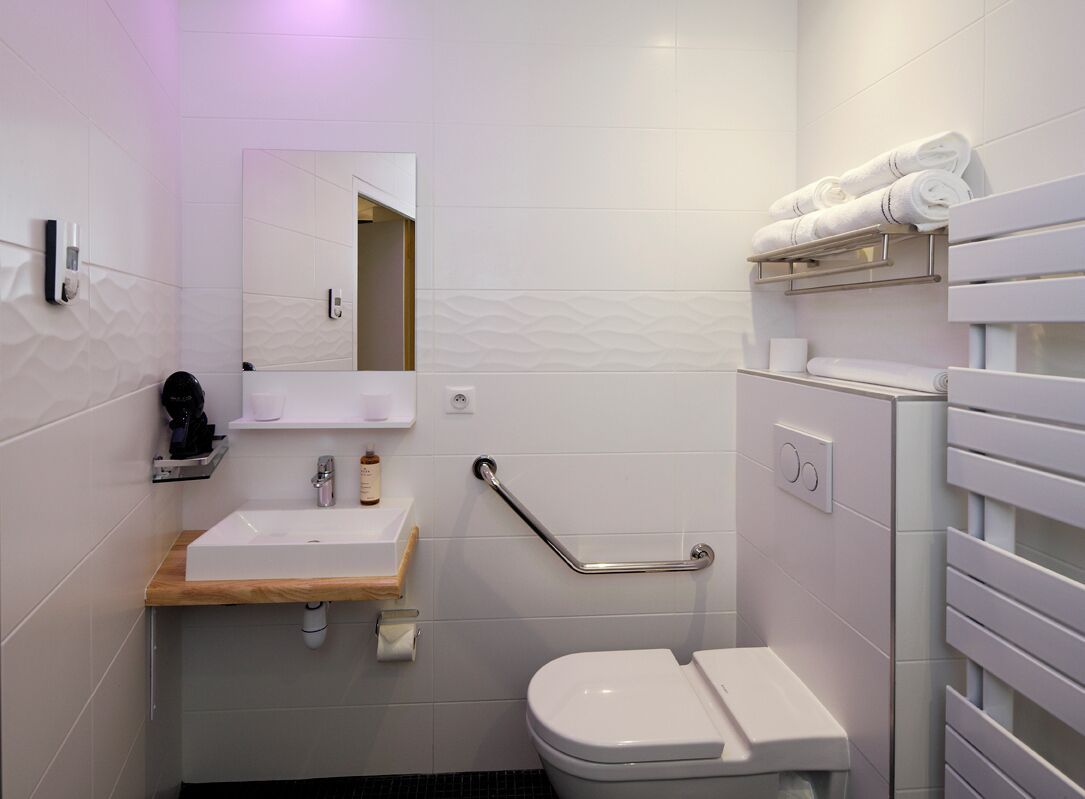 Un baño moderno con lavamanos, espejo y retrete blancos. Hay un perchero en la pared y un pasamanos para facilitar el acceso.