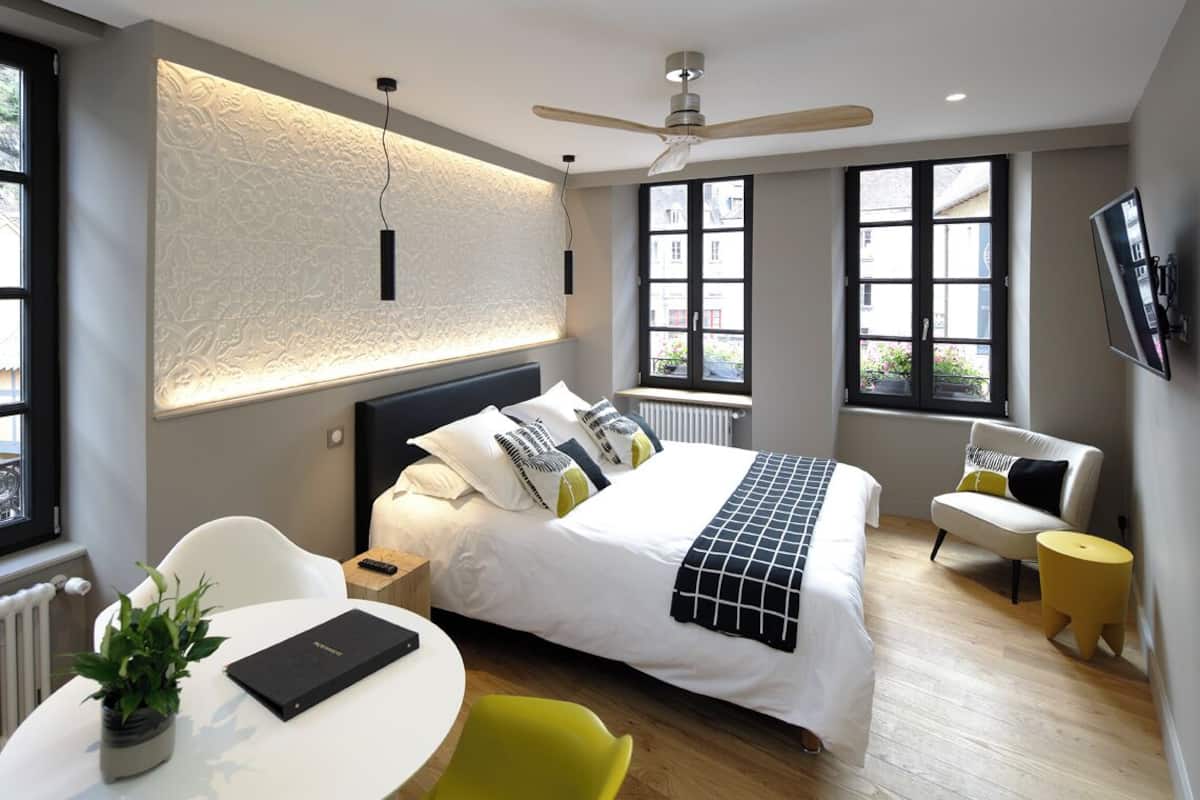 A modern bedroom with a large bed, a ceiling fan, a window with a view, a small table with a plant, and a TV mounted on the wall.