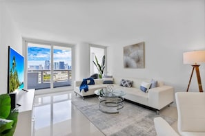 A modern living room with a white sofa, a glass coffee table, and a large window offering a city view. - Downtown Seaclusion (Miami)