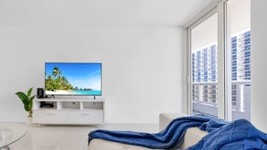 Ein modernes Wohnzimmer mit einem großen Flachbildfernseher auf einer weißen Unterhaltungsanlage, einem beige farbenen Sofa und einem großen Fenster mit Blick auf ein Gebäude.