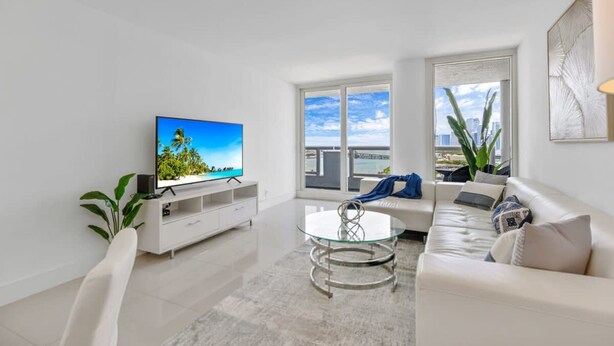 Ein modernes Wohnzimmer mit Flachbildfernseher, Glastisch, weißem Sofa und großem Fenster mit Blick auf die Stadt.